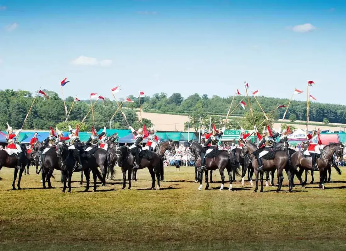 A group of uniformed cavalry riders on horseback, holding lances with red and white flags, standing in an open field.