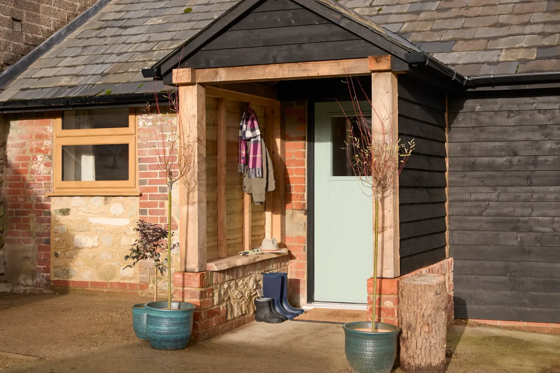 A wooden porch entrance with a light green door, flanked by brick and stone walls, a dark timber side, and two potted trees.