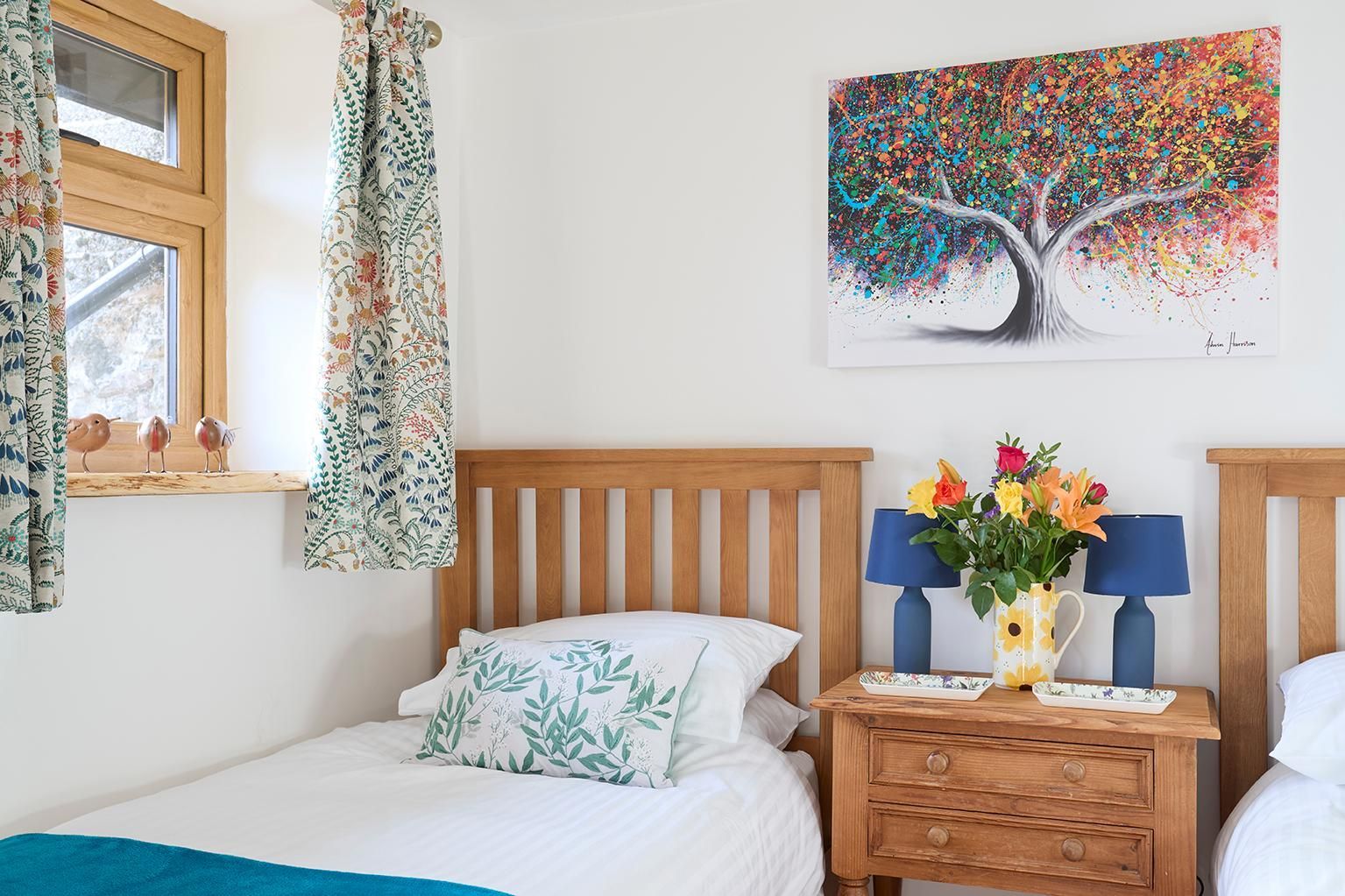 Two twin beds with wooden headboards, a bedside table with a floral vase, and a colorful tree painting on a white wall.
