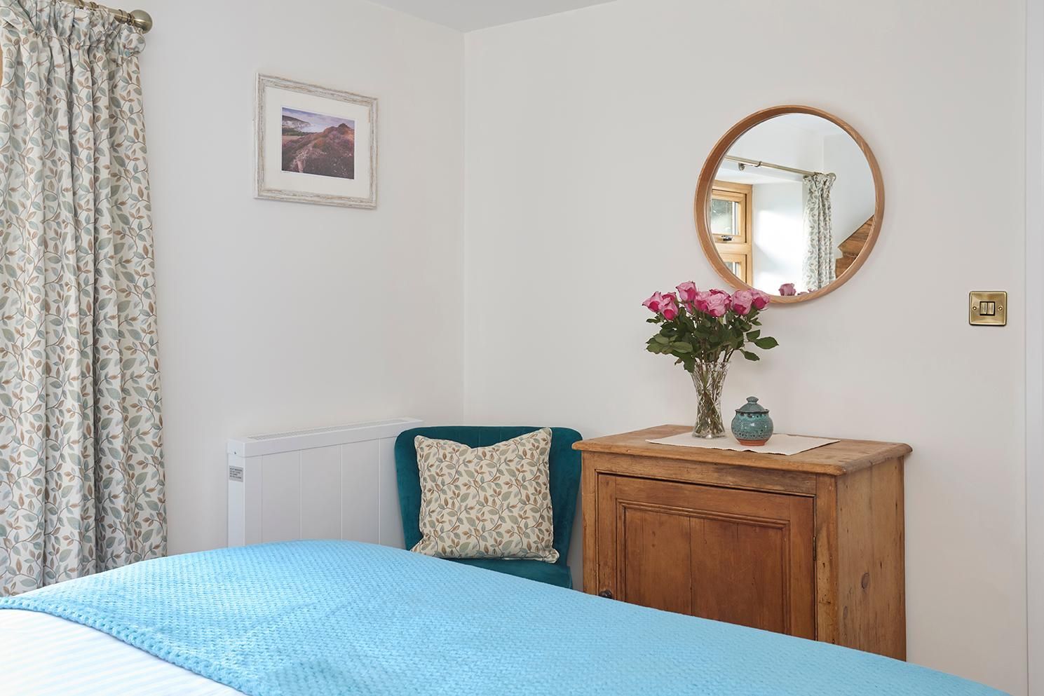 A bedroom corner with a teal armchair, a blue bedspread, a wooden cabinet, a mirror, and a floral-patterned curtain.