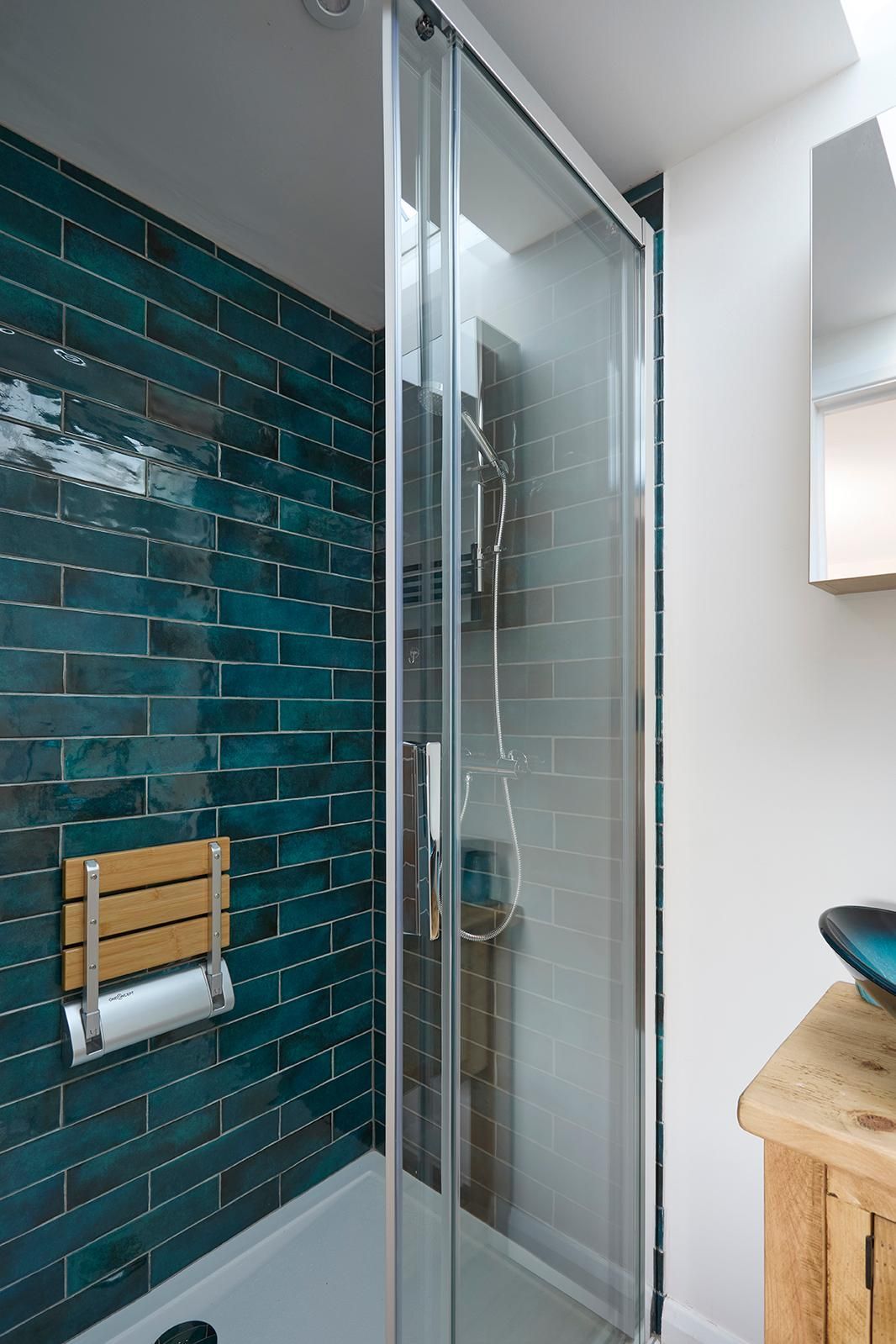 A walk-in shower with dark teal subway tiles and a wooden fold-down seat, next to a rustic wooden vanity and sink.