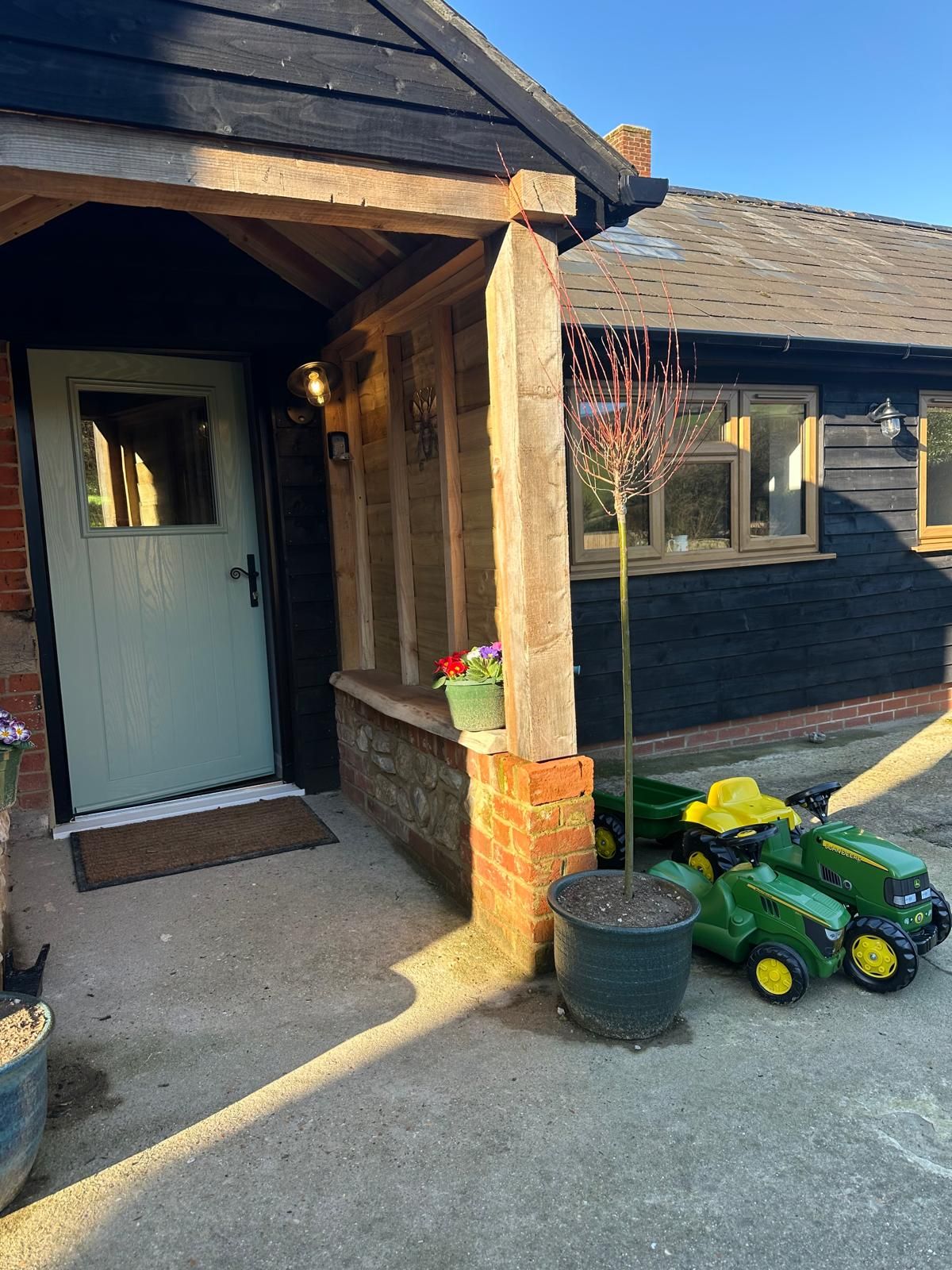 A light grey door under a timber-framed porch with a small tree in a pot and two green toy tractors on a paved driveway.