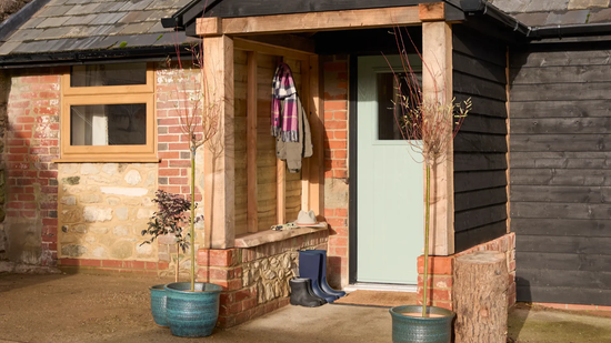 A wooden porch shelter covers a light green door of a rustic building with brick walls and black timber siding.