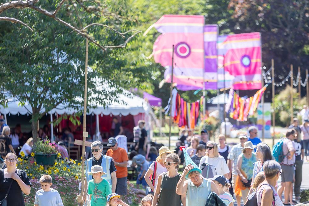A crowd walks past colorful banners and a white event tent at an outdoor festival on a sunny day.