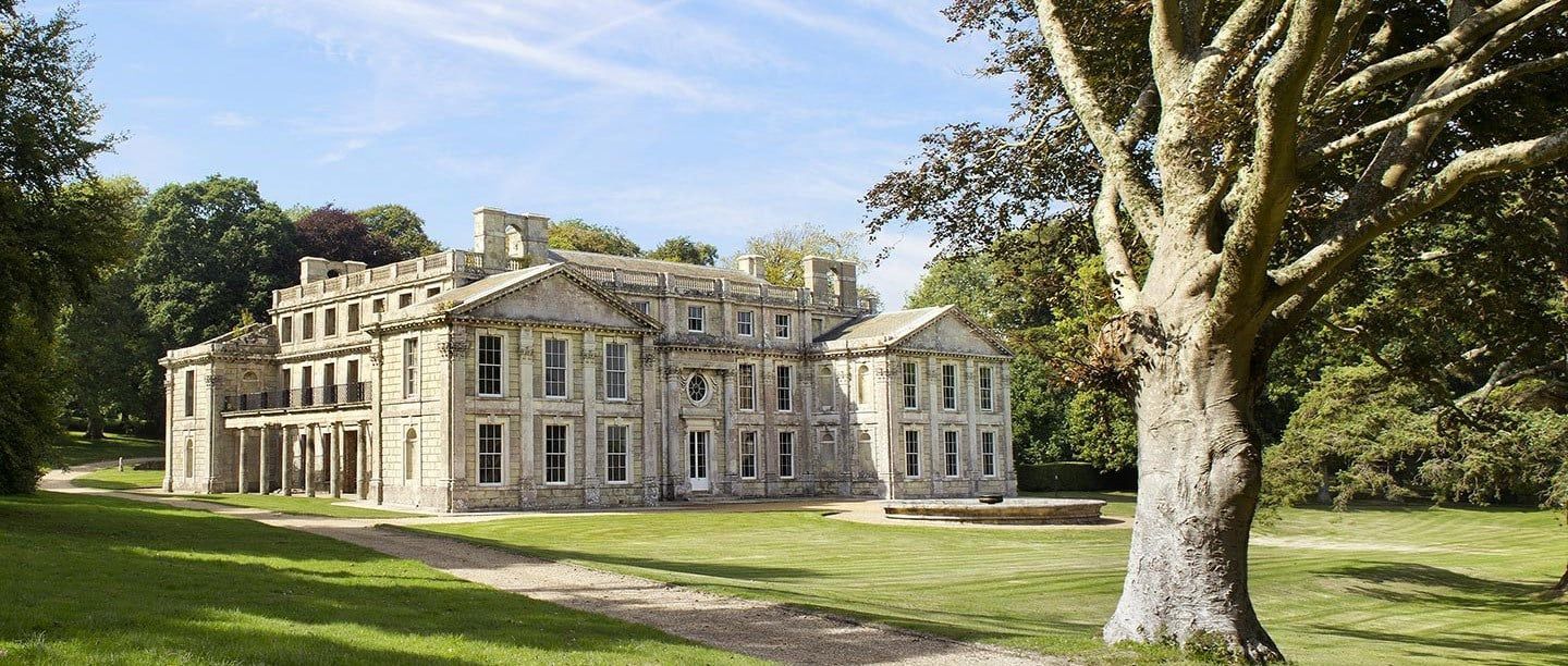 A large, historic stone mansion with classical architecture stands in a grassy parkland near a large tree under a blue sky.