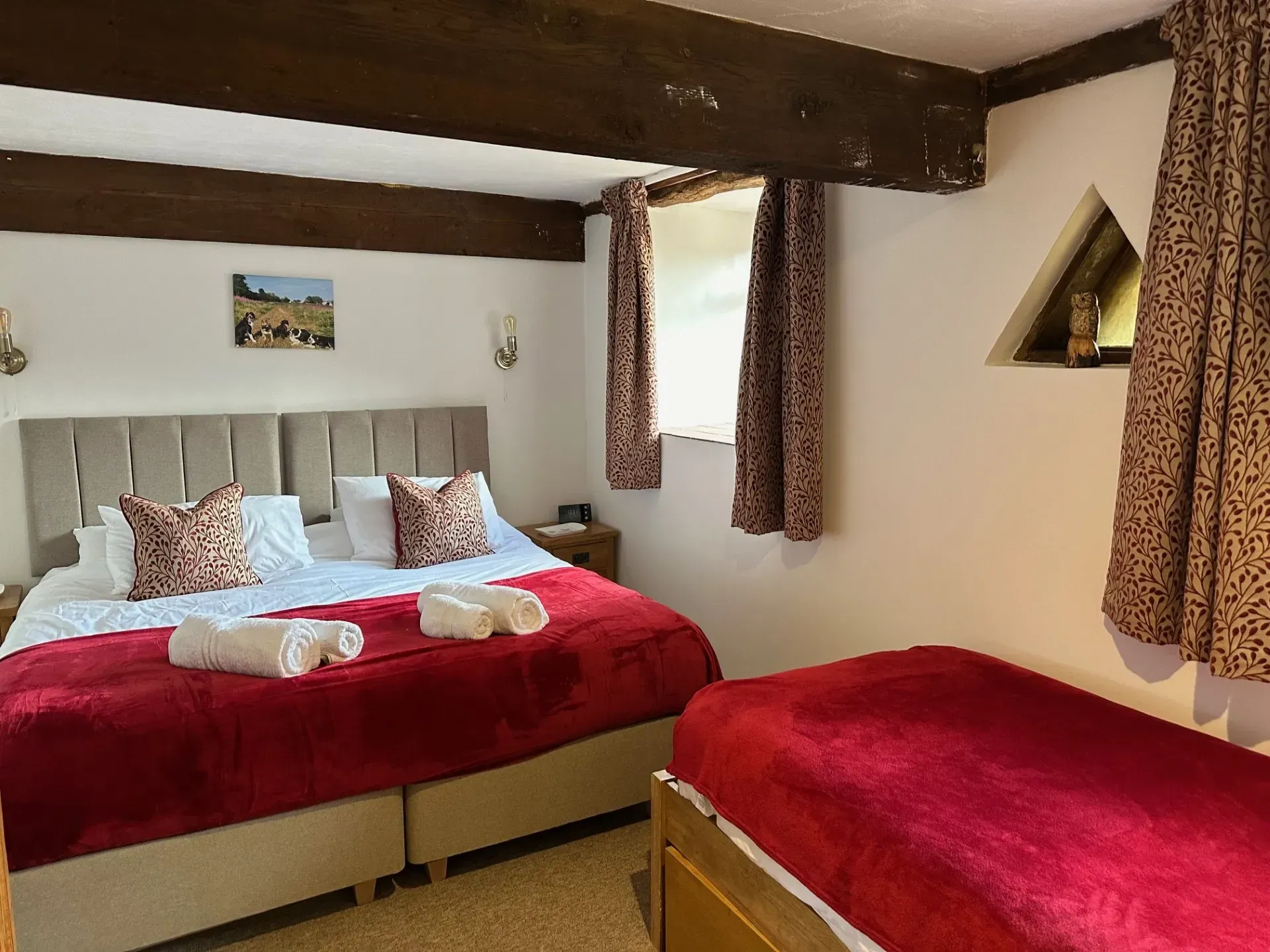 A bedroom with a large bed and a smaller bed, both featuring red blankets, wooden ceiling beams, and patterned curtains.