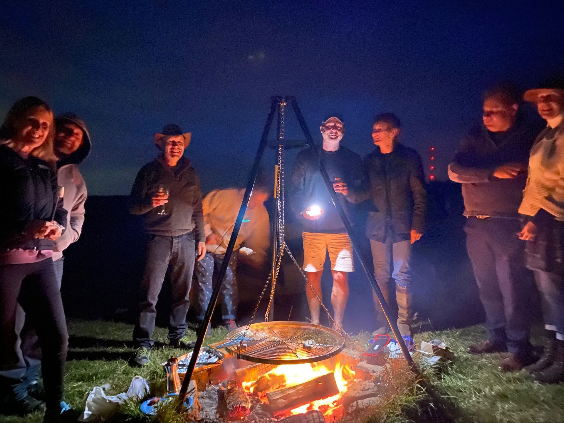 Group gathered around a glowing campfire at night, with a tripod cooking pot in an outdoor setting