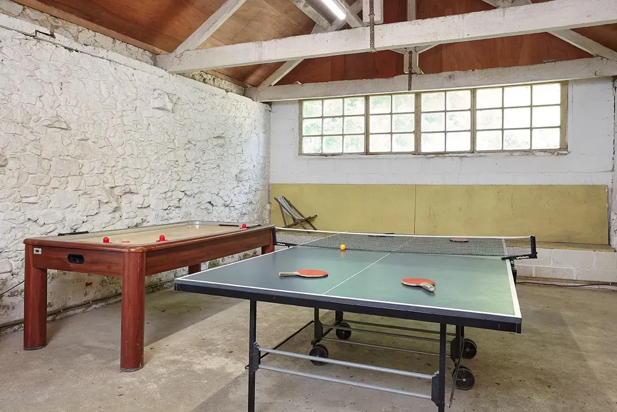 A game room featuring a green ping pong table and a brown air hockey table in a rustic room with white, textured walls.
