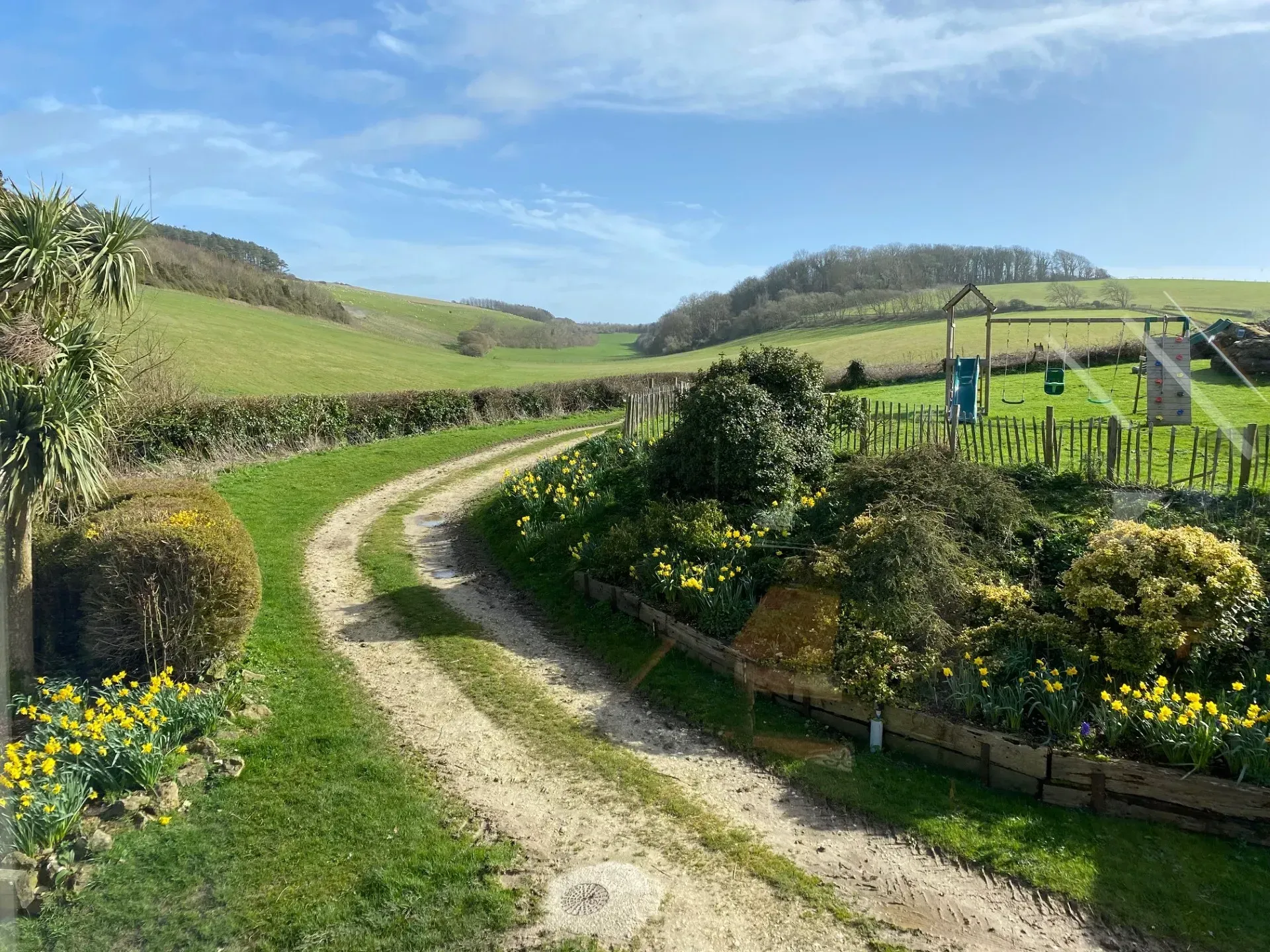 A dirt path curves through a sunny, rolling green landscape with a garden, flowering plants, and a wooden playground set.
