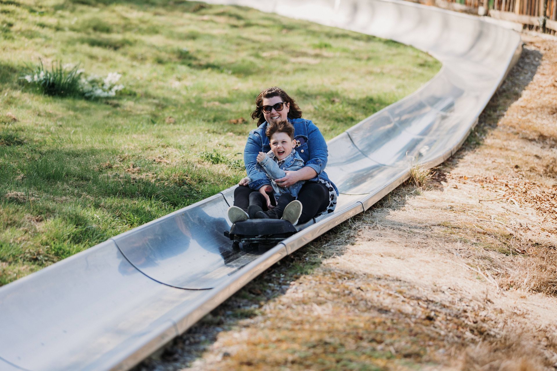 Two people smiling as they ride a metal bobsled track through a grassy field.