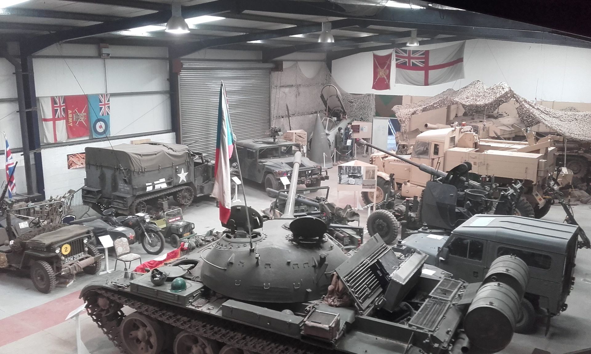 A wide-angle interior view of a military museum hangar featuring a tank, various historic vehicles, and flags on the walls.
