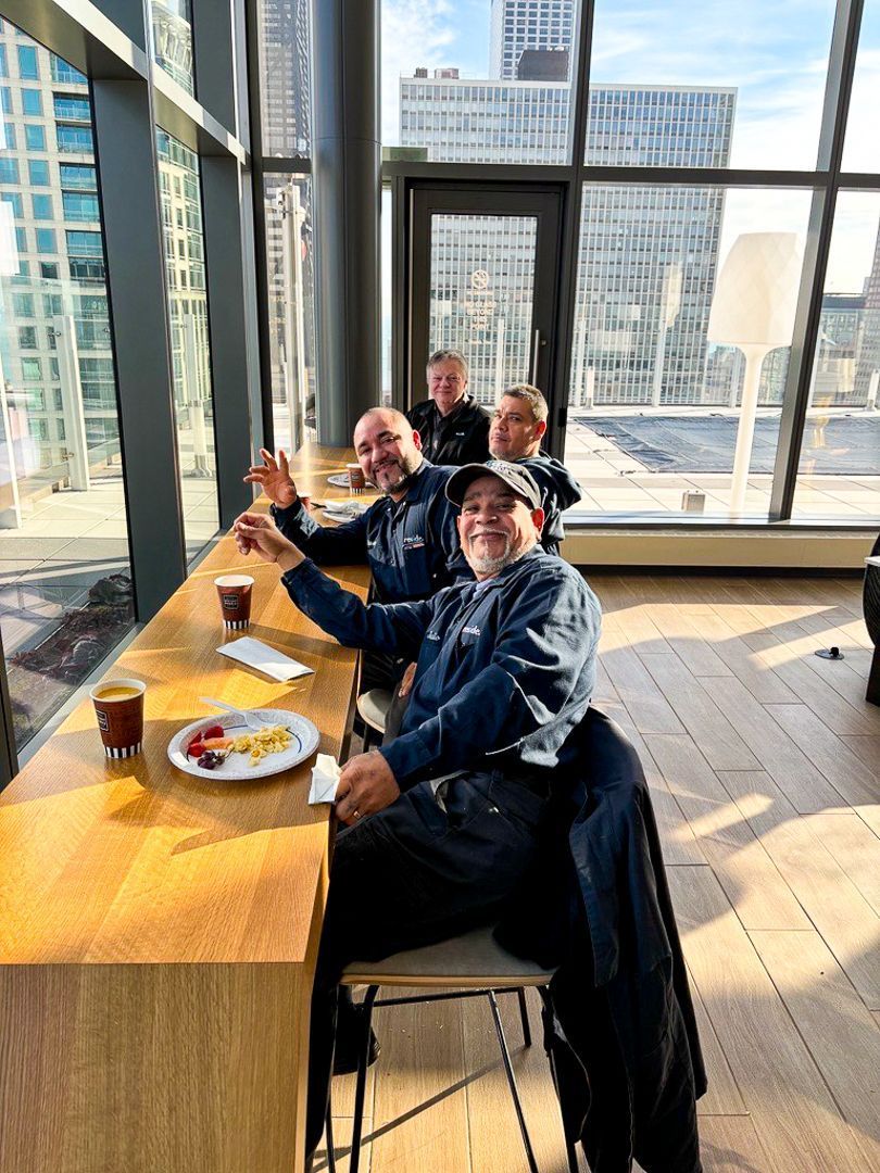 People seated at a sunlit table in a city office lounge, toasting with drinks near large windows.