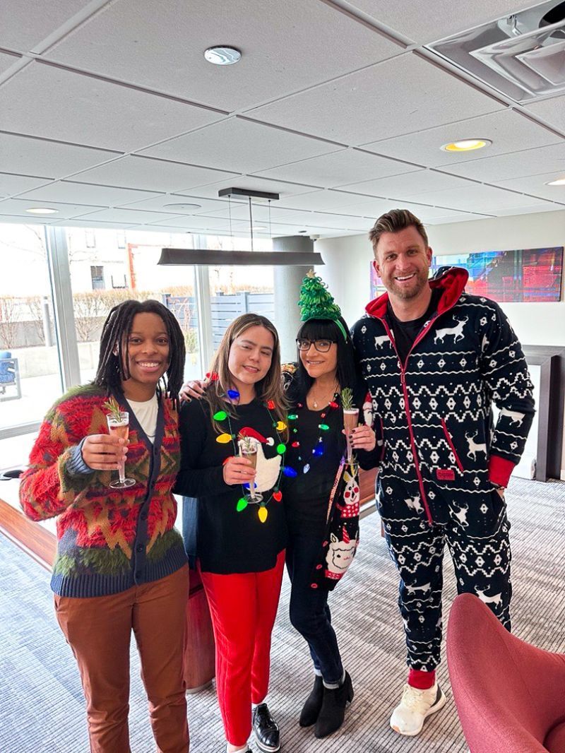Four people in festive holiday sweaters pose indoors near a decorated tree.