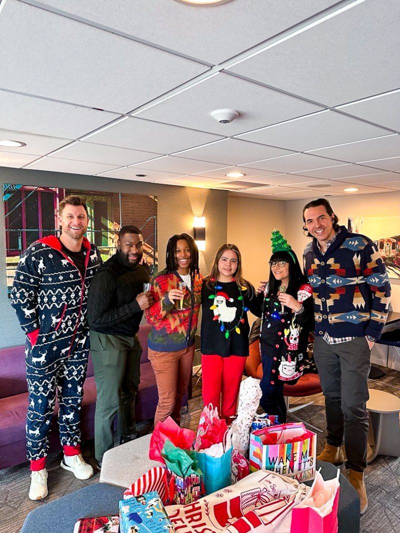 People in holiday sweaters pose by a table of wrapped gifts in a festive indoor room.