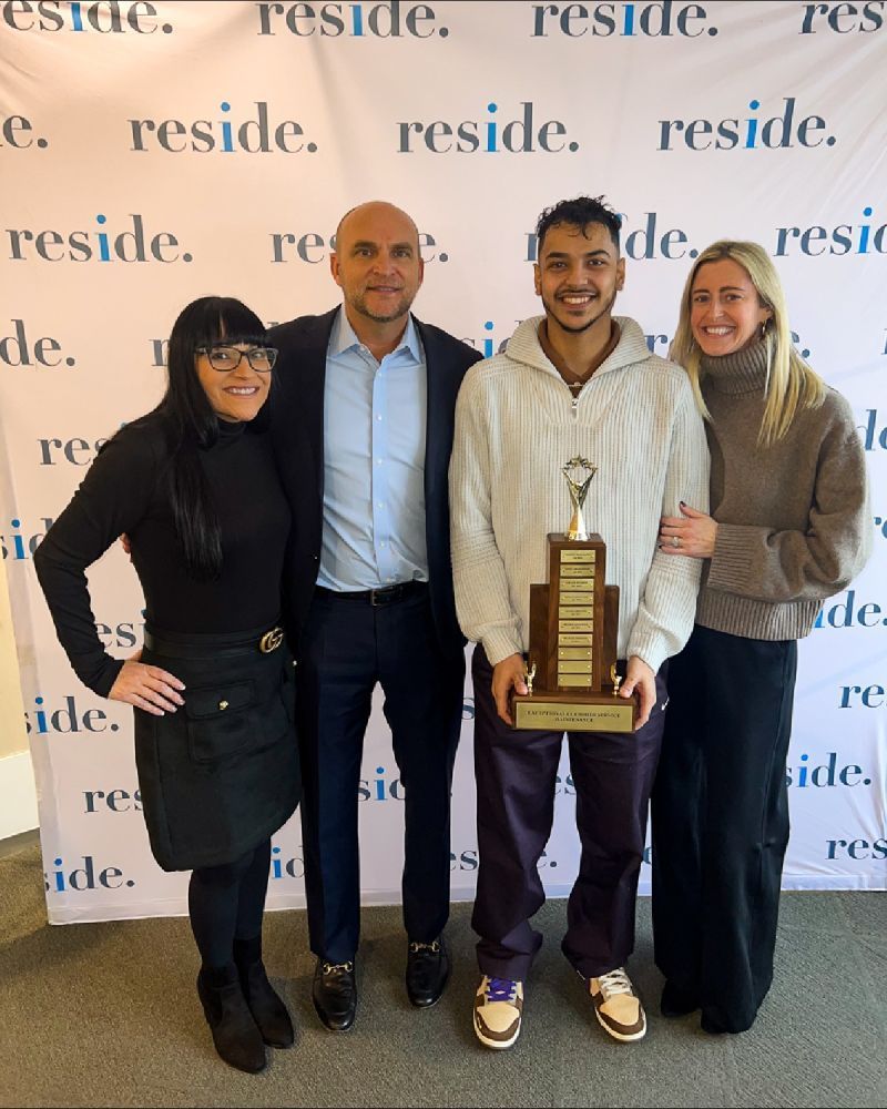 Four people posing indoors, one holding a tall trophy, in front of a repeating “reside” backdrop