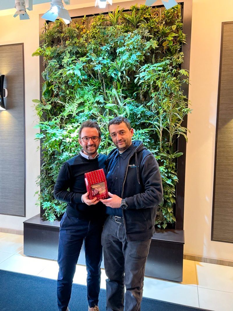Two men standing indoors holding a red award in front of a green plant wall.