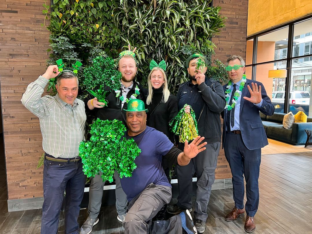 Group of six people posing indoors with green pom-poms and festive props, smiling in a lounge area.