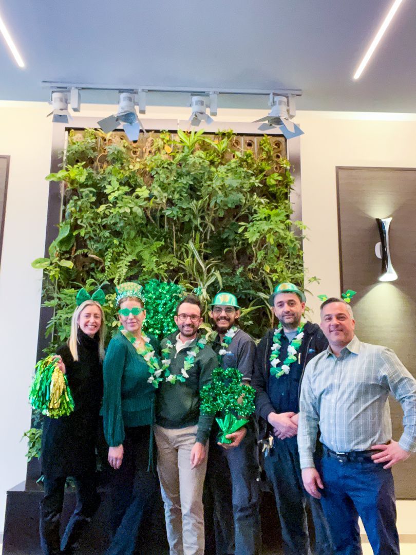 Five people posing in front of a green plant wall indoors, wearing green leis and smiling.
