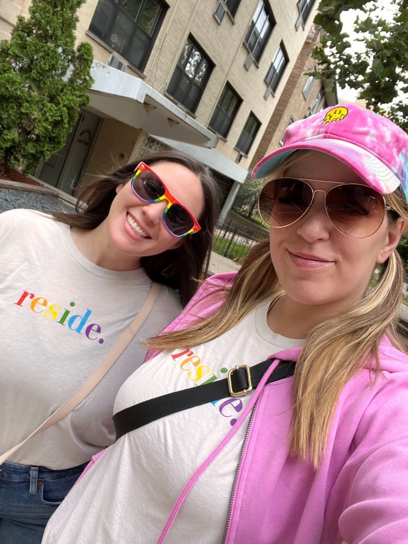 Two women smiling outdoors, one in a pink cap and jacket, the other in a white shirt and sunglasses.