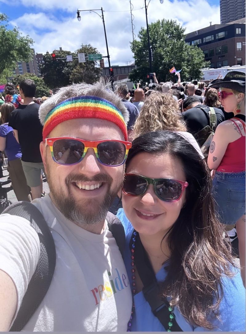 Two people smiling for a selfie at a sunny outdoor street event, one wearing a rainbow beanie and red sunglasses