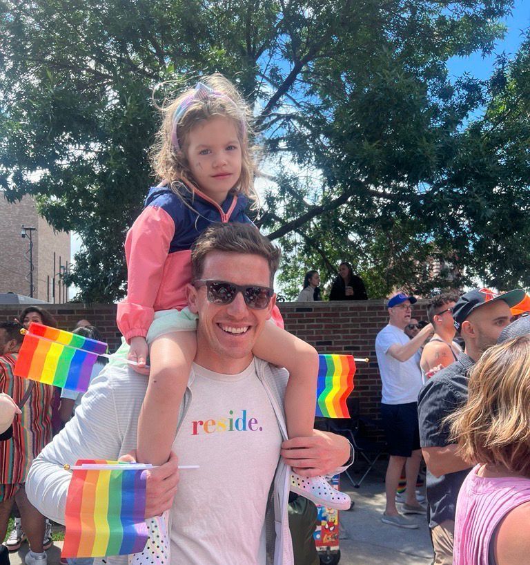 Adult carrying a child at a rainbow parade, both smiling amid Pride flags and a sunny crowd.