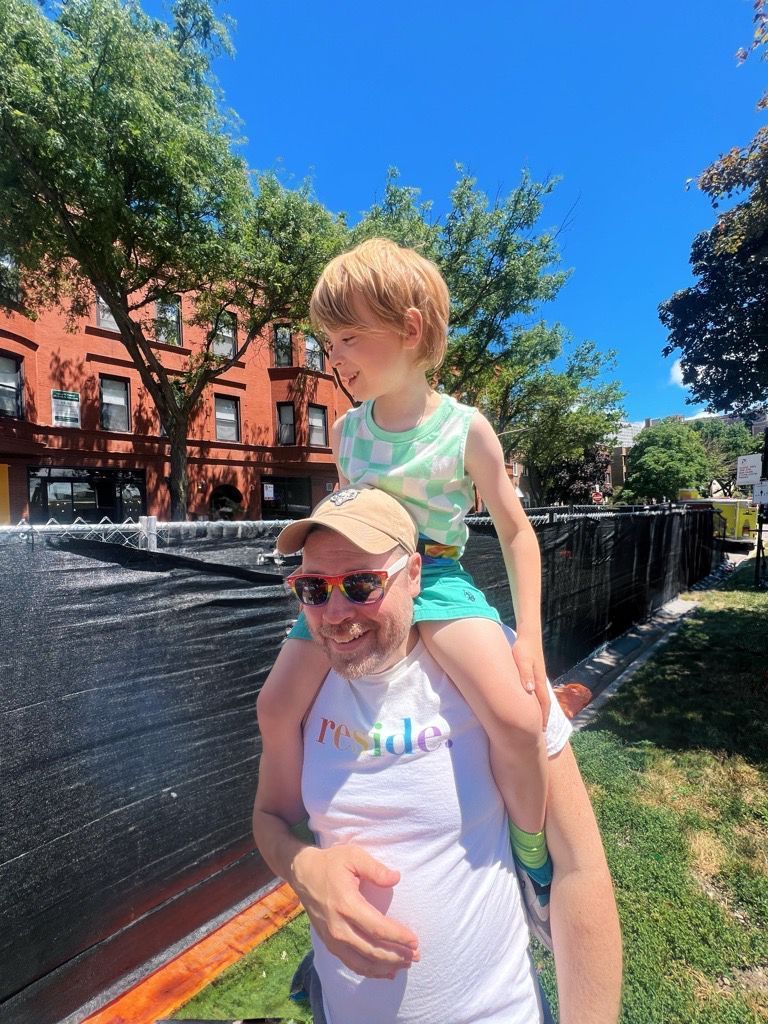 Adult carrying child on shoulders beside a black fence on a sunny street with brick buildings and trees