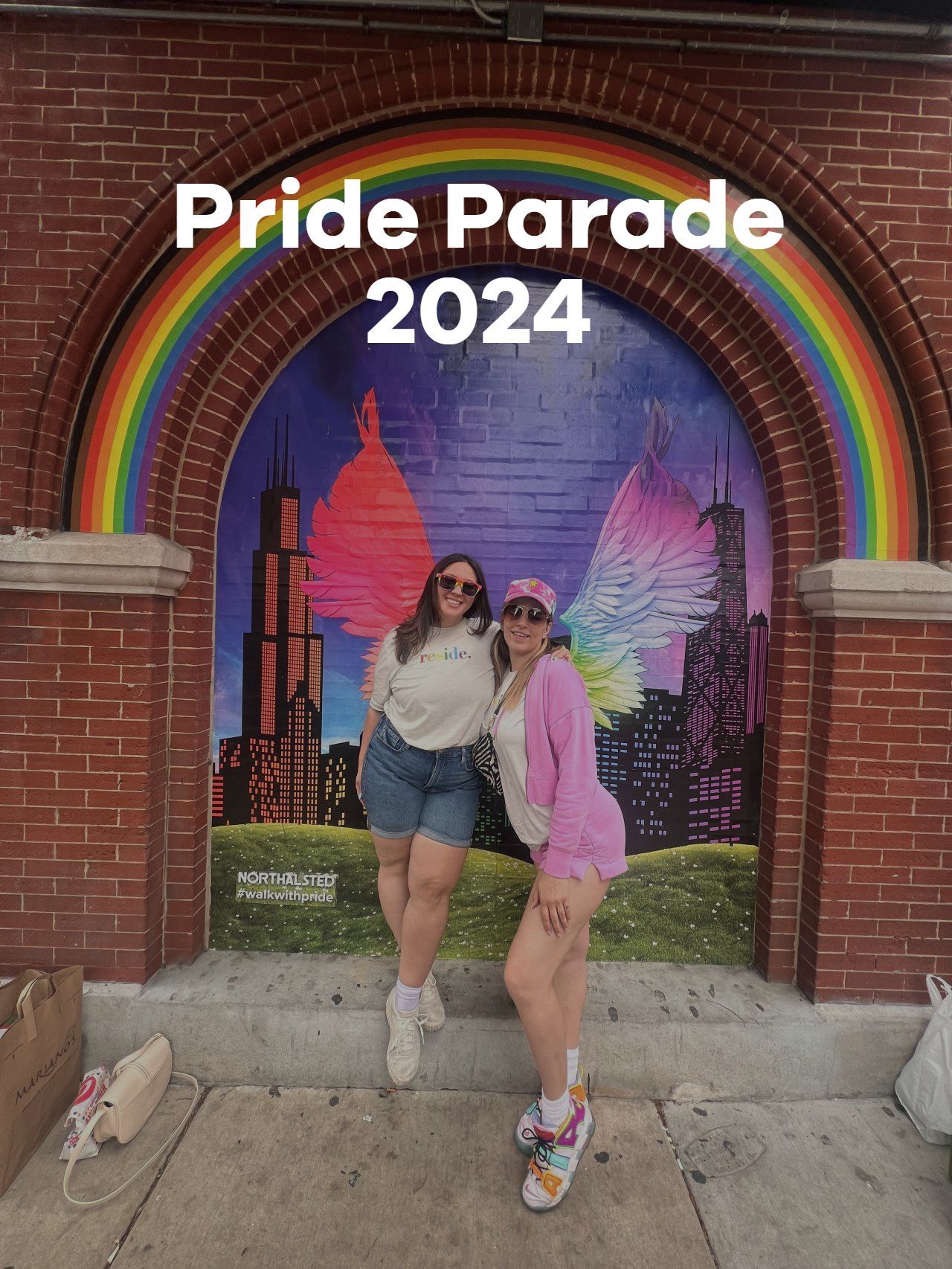 Two people posing under a rainbow arch at a Pride Parade 2024 mural with a city skyline backdrop
