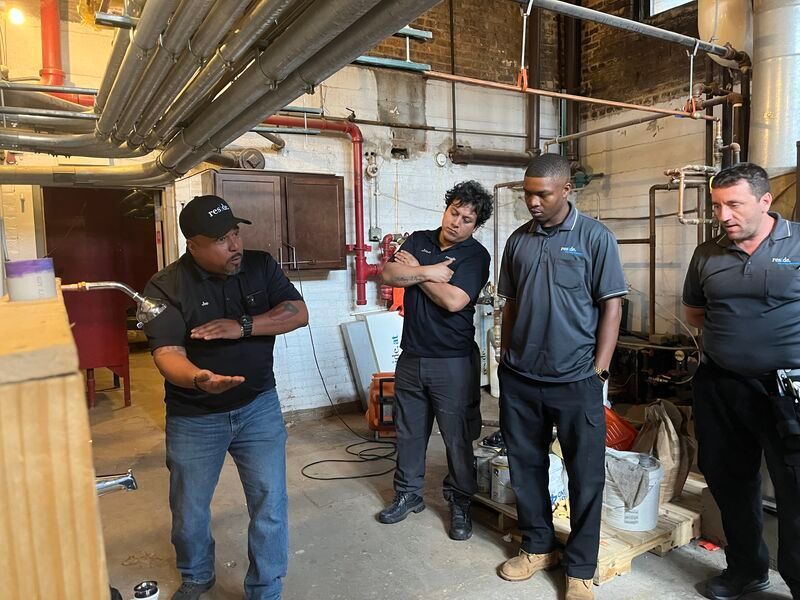 Four workers stand in an industrial room inspecting equipment under overhead pipes.