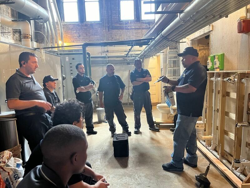 Workers gathered in a warehouse aisle, listening to a man speaking beside stacked shelves and boxes.