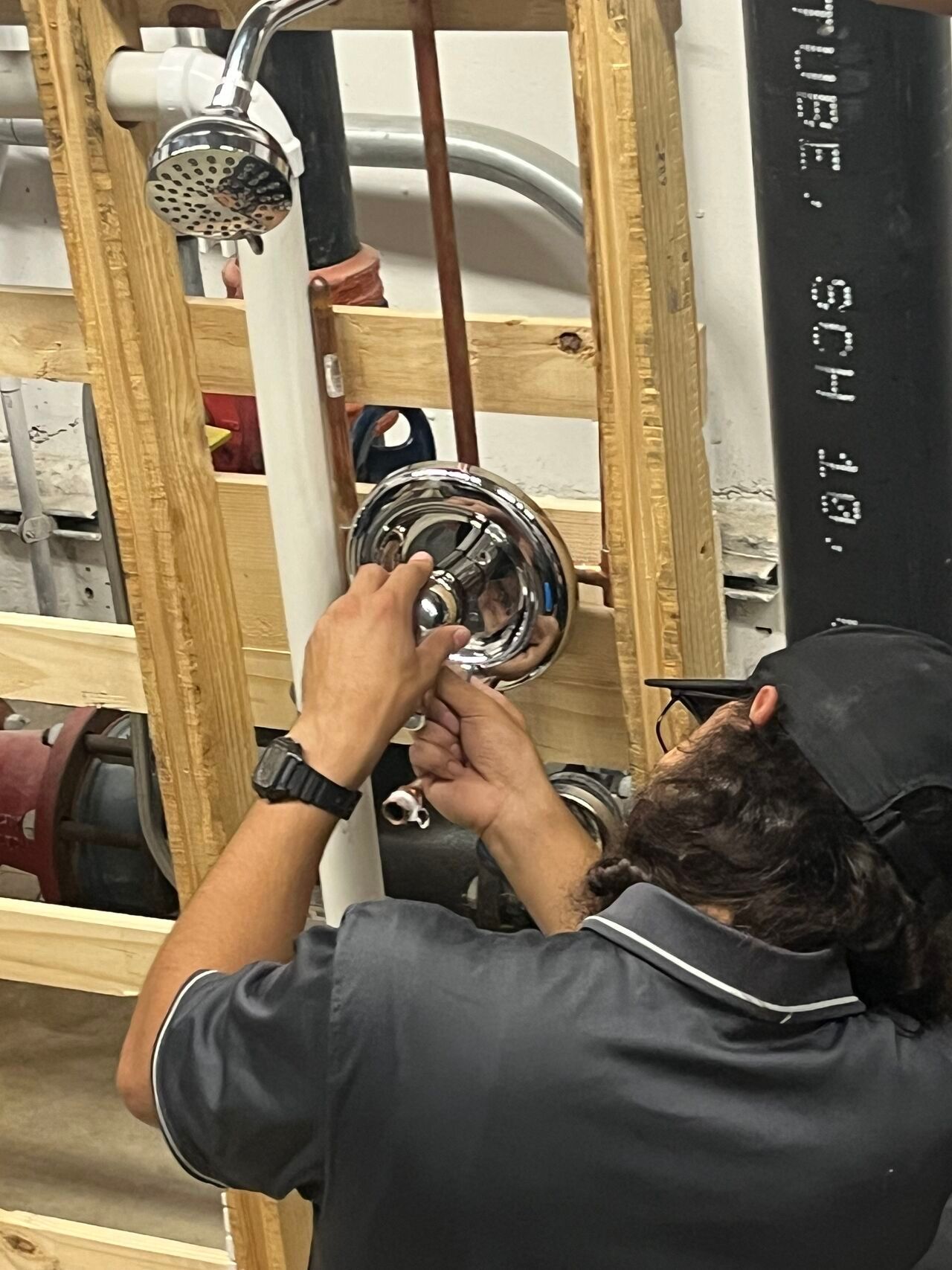 Plumber installing a chrome shower fixture on exposed bathroom plumbing with wooden framing and black pipes