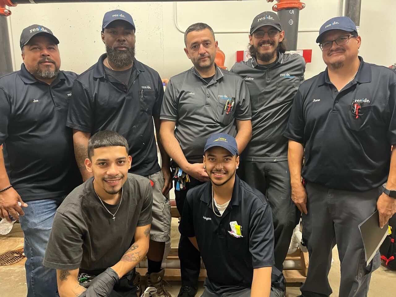 Seven people in matching black shirts and caps pose together indoors, smiling at the camera.