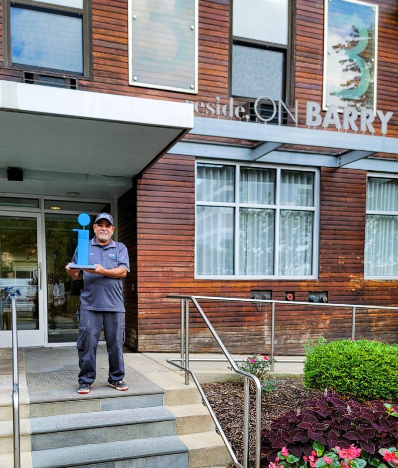 Man standing on steps outside a brick building with a sign reading “On Bakery,” holding papers.