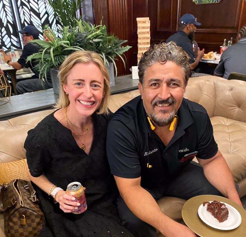 Two people smiling on a beige couch in a lounge, holding a drink and dessert plate.