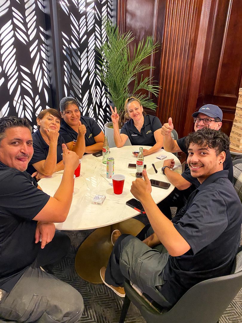 Five friends clink drinks around a white table in a cozy restaurant with wood walls and plants.