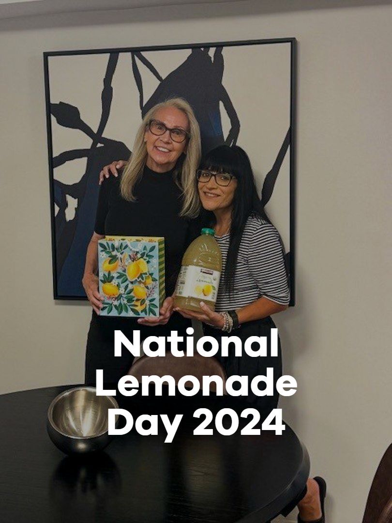 Two people holding lemonade stand signs in front of a wall art display, with “National Lemonade Day 2024” text.