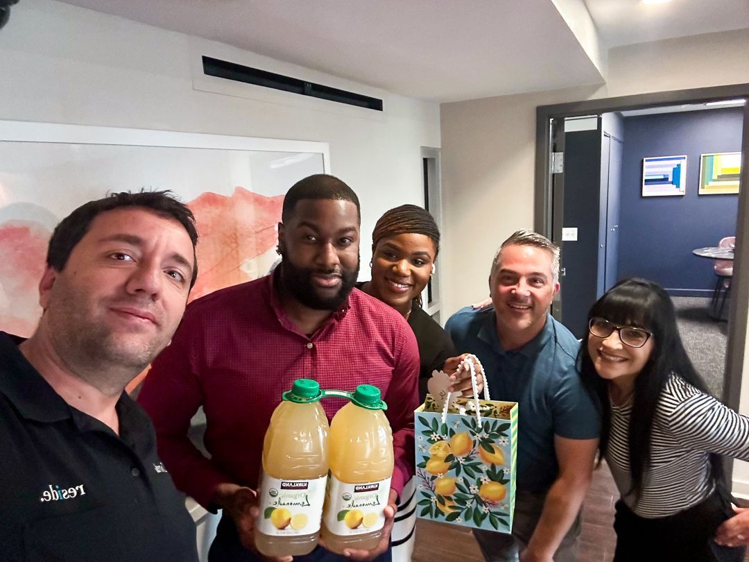 Group of five people posing indoors with orange juice bottles and a floral gift bag