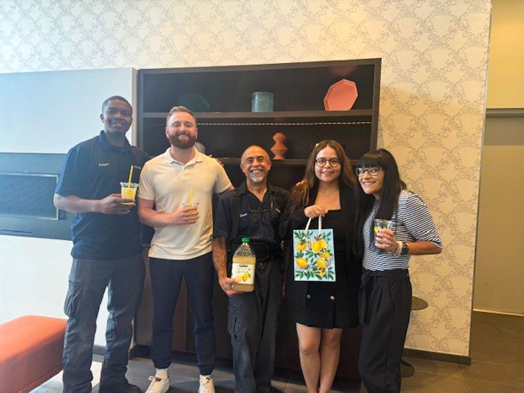Five people posing indoors, holding drinks and a bouquet, in front of a shelf and patterned wall.