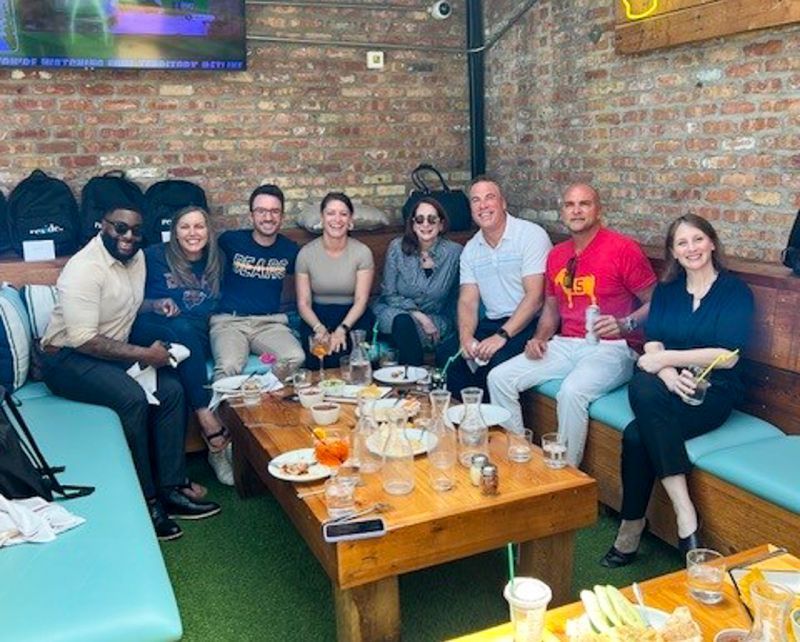 Group of friends seated on a patio around a table with drinks, chatting against a brick wall.