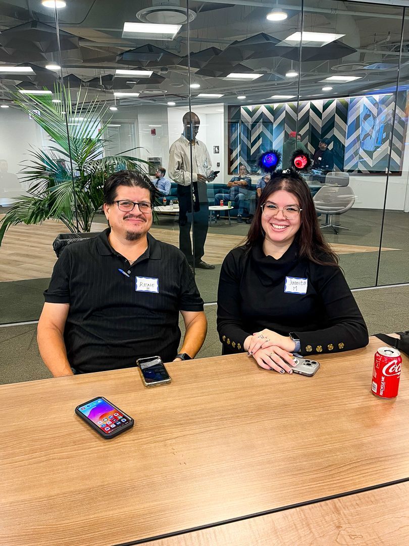 Two people seated at a conference table in an office, smiling with drinks and a phone in front of them.