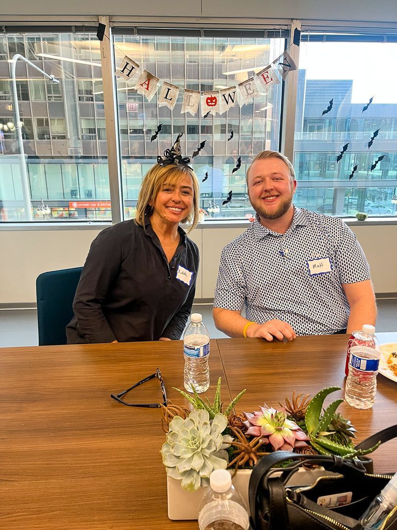 Two people smiling at a conference table in a bright office, with flowers and drinks in the foreground.
