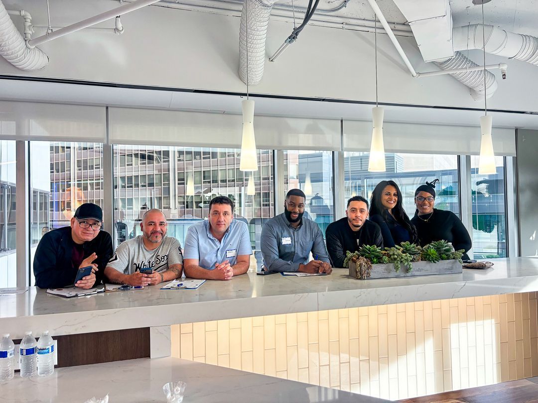 Six people lean on a counter in a bright modern office kitchen, smiling at the camera.