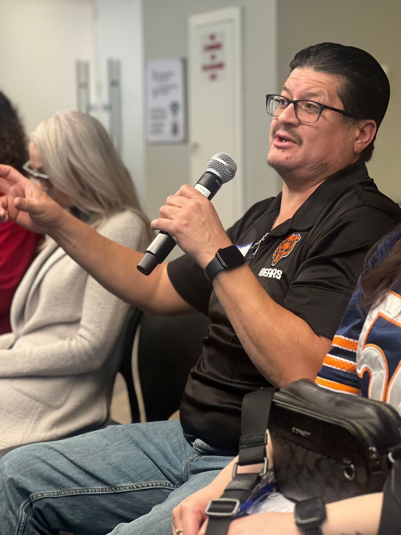 Man speaking into a microphone at a meeting, gesturing while seated beside others in a conference room.