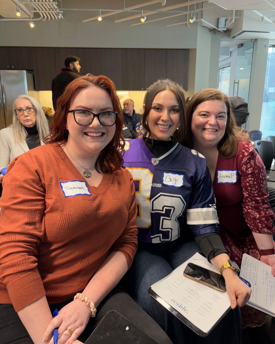 Three smiling people seated together at an indoor event, one holding a notebook.
