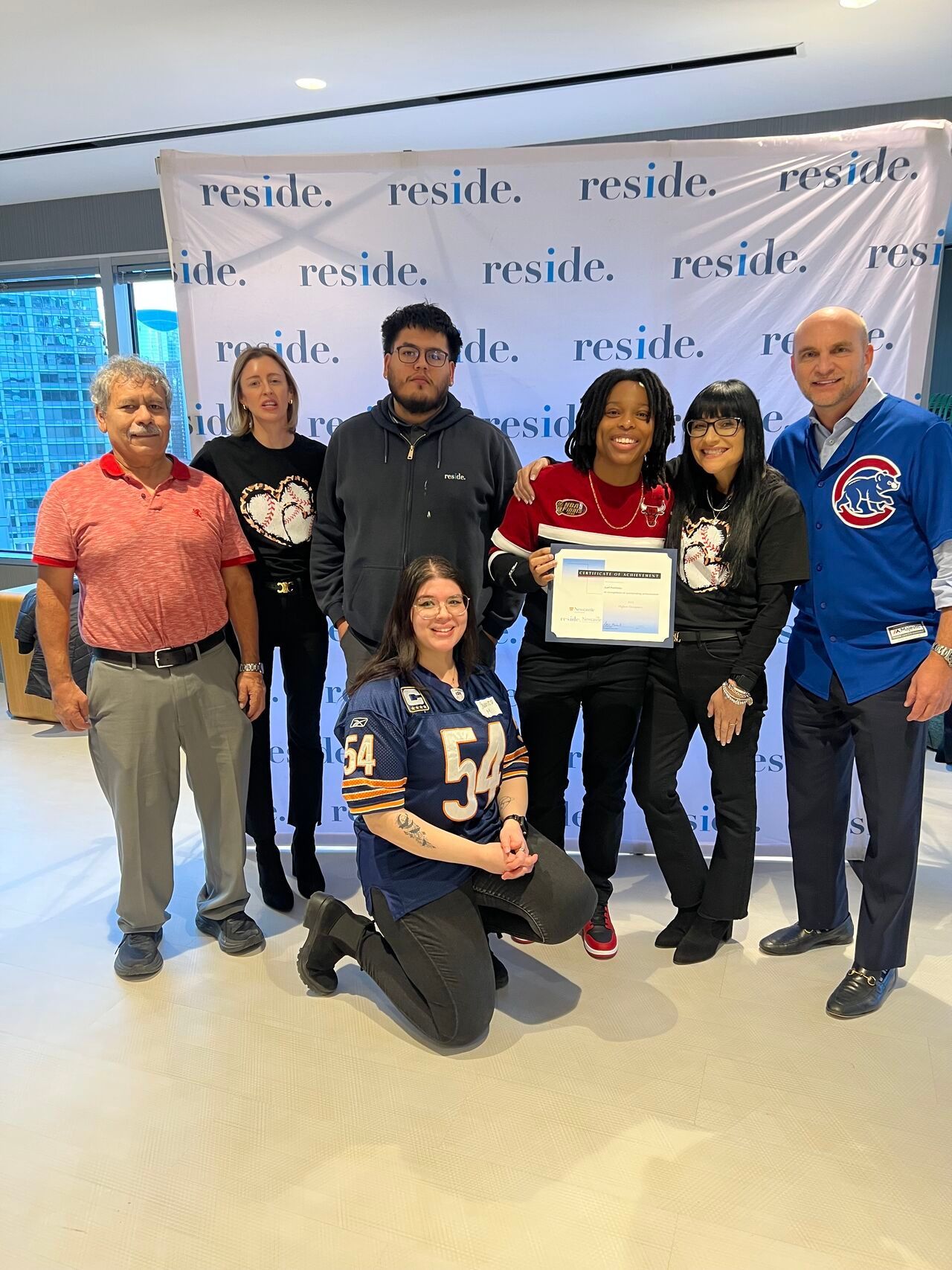 Group of seven posing indoors, one holding a certificate, in front of a repeated “reside” backdrop