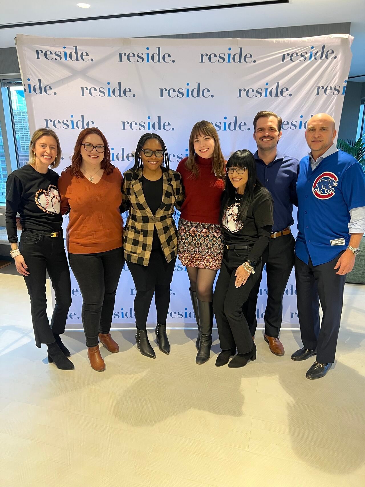 Seven people posing together at an indoor event with a branded backdrop, smiling at the camera.