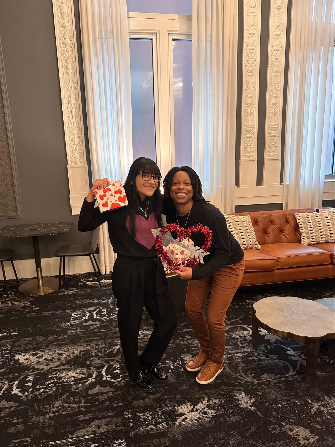 Two women posing indoors, smiling with bouquets in a elegant lobby with tall windows and patterned carpet
