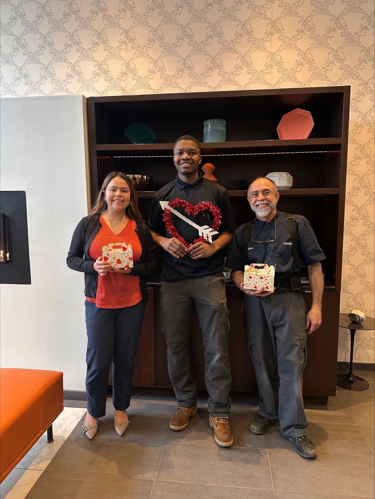 Three people standing indoors holding small boxes in front of a dark bookshelf.