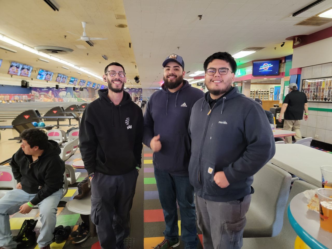 Three friends standing in a bowling alley, smiling near the lanes and seating area.
