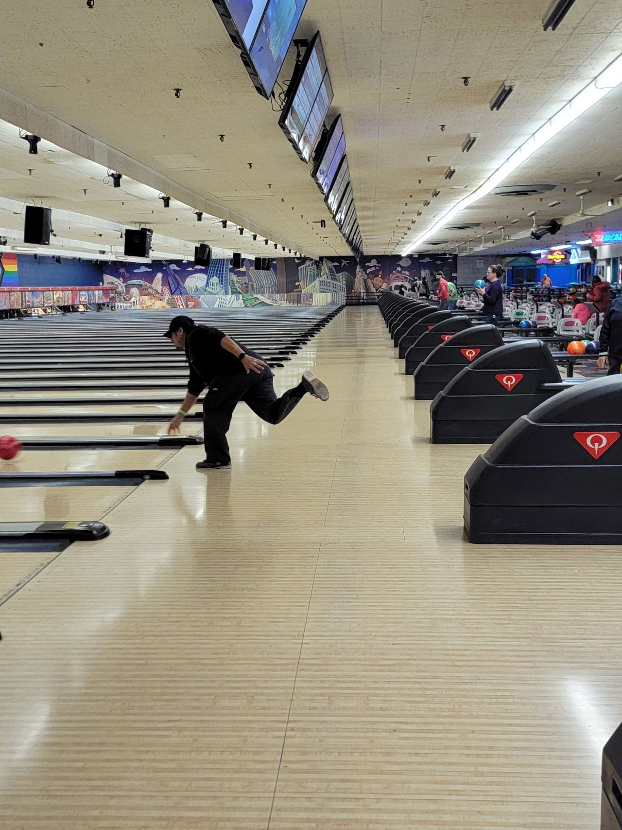Bowler mid-release on a bowling lane in a brightly lit alley, with rows of lanes and ball returns visible