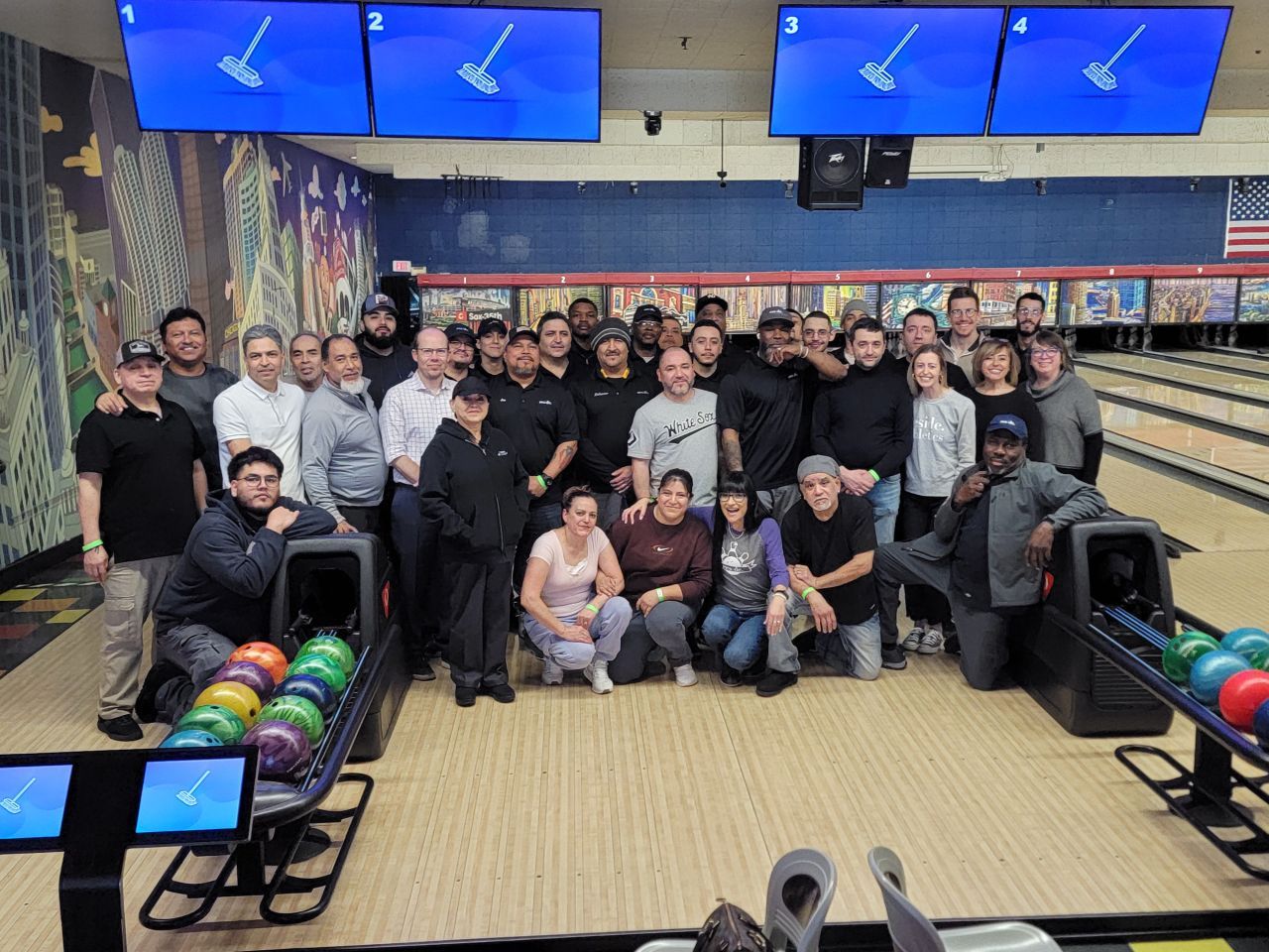 Group of people posing in a bowling alley with lanes, pins, and blue overhead screens.